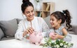 © Mrityunjoy - Mother and Daughter Bonding Over Saving with Pink Piggy Bank at Home, Smiling and Engaged in Playful Activity