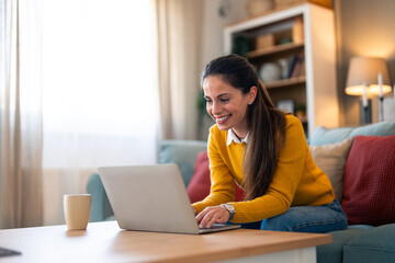 Wall Mural - Cheerful female in a yellow sweater engages in an online chat, seated comfortably on a couch with her laptop in a bright living room setting.