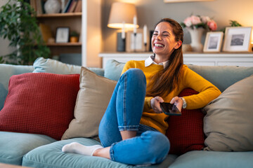 Wall Mural - A cheerful Caucasian woman wearing a cozy yellow sweater smiles brightly as she interacts with her smartphone, comfortably seated on a couch in a homey living room setting.