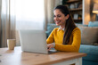 © Dorde - A joyful Caucasian woman engages in a video call on her laptop, seated comfortably on the floor by her living room table, dressed in casual attire.