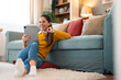 © Dorde - A cheerful woman engages in a video call using her tablet, comfortably seated on the floor by the couch in a cozy living room, with coffee in hand.