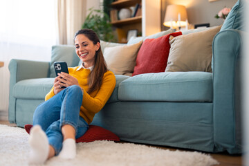 Wall Mural - A cheerful woman enjoys a video call on her smartphone, seated comfortably on the floor by the couch in a cozy living room.