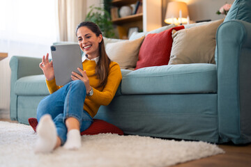 Wall Mural - A cheerful woman enjoys a digital connection at home, engaging through video chat on her tablet while sipping coffee, seated comfortably by her couch.