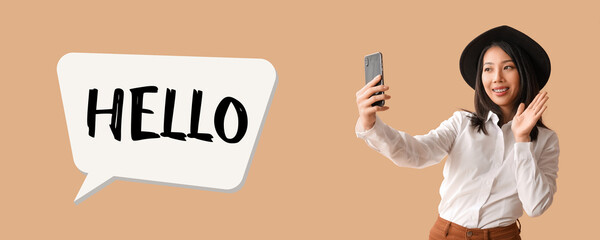 Poster - Beautiful young Asian woman with mobile phone waving hand on beige background