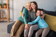 © Dorde - A joyful grandmother, her daughter, and granddaughter share a snug hug on a cozy sofa, radiating happiness in a well-lit living room.