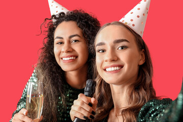 Poster - Beautiful young woman in party hats with bottle of champagne celebrating Birthday and taking selfie on red background