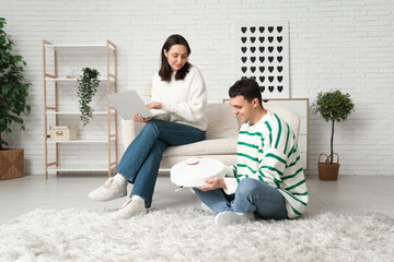 Poster - Young couple with modern robot vacuum cleaner and laptop in living room