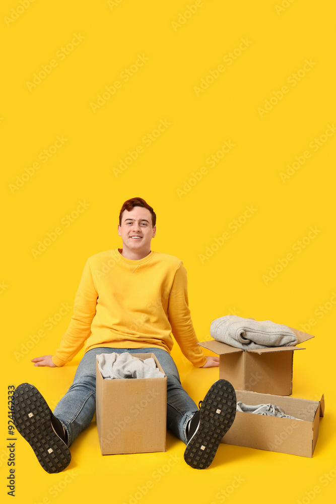 Happy young man sitting near wardrobe boxes with clothes on yellow background