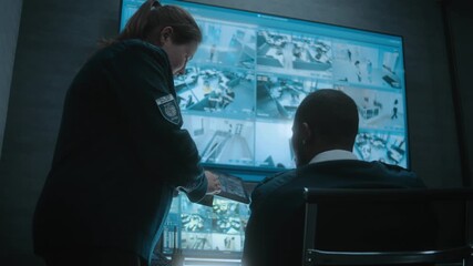 Female worker shows surveillance footage to African American security officer using tablet computer. Multiethnic security operators monitor CCTV cameras on computer monitors and big digital screen.