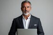 © Dream Canvas - Serious handsome latin older man in formal suit holding desktop computer. Portrait of focused middle age mature businessman using pc laptop for business work. Isolated white background, Generative AI