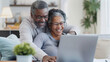 © ifoto - Black senior couple smiling while using a laptop at home, sitting in their living room