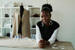 © pressmaster - Young African American female tailor or fashion designer looking at camera while standing by table with unrolled grey textile