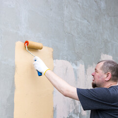 Wall Mural - A man wearing gloves paints a wall with a roller brush. The wall is gray, and he is applying a coat of tan paint. The sun casts a warm glow on the wall and the man's face.