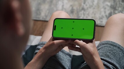 Poster - caucasian teenager holds mobile phone with green screen, watches video or movie sitting on sofa. you