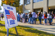 © CozyDigital - Vote here. Sign with American flag next to polling place. A line of people waiting to vote.