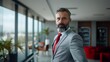 © Scott - A confident businessman with a well-groomed beard, wearing a gray suit and red tie, standing in a stylish office with large windows.