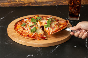 Delicious and appetizing slice of pizza served on a wooden cutting board topped with various ingredients such as cheese, pepperoni, and fresh herbs, with a hand is about to cut into the pizza.