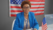 © Krakenimages.com - Confident african american woman wearing glasses and blue blazer sitting in front of an american flag in an indoor setting.