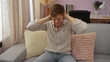 © Krakenimages.com - Young man sitting on couch at home covering his ears in frustration while inside a cozy and well-lit living room with pillows and bookshelves in the background