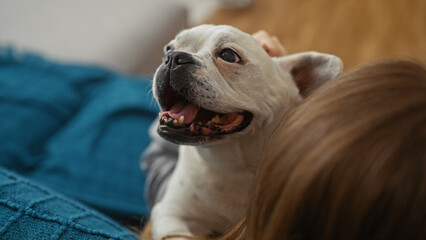 A young blonde woman in a living room holds her happy canine pet, a cute white dog, while sitting indoors in an apartment.