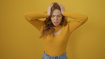 Wall Mural - A young beautiful blonde caucasian woman in a yellow sweater stands over a yellow background, appearing stressed and isolated.