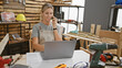 © Krakenimages.com - Young caucasian woman talks on phone at a carpentry workshop with laptop and tools.