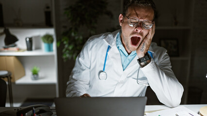 Canvas Print - Exhausted young man, a doctor wearing glasses, yawns intensely in a clinic room with a computer and plants in the background.