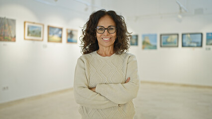 confident middle-aged hispanic woman with glasses standing with arms crossed in an art gallery, disp