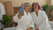 © Krakenimages.com - Mature women enjoying a spa day indoors, wearing robes and taking a selfie, surrounded by a relaxing wellness interior