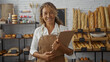 © Krakenimages.com - Woman standing in a bakery room wearing an apron and holding a clipboard, surrounded by various breads, baguettes, and pastries on shelves.