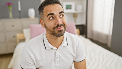 Sticker - Young hispanic man with beard in casual attire looking up thoughtfully, positioned indoors in a modern bedroom setting.