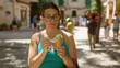 © Krakenimages.com - Young hispanic woman enjoying ice cream outdoors in valldemossa, mallorca, spain, wearing glasses and a green top, with a sunny street setting in the background.
