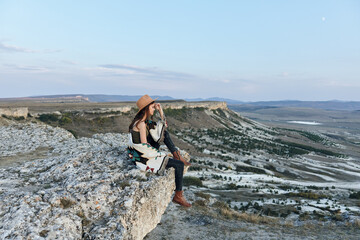 Wall Mural - Serene woman in hat sitting on rock, gazing at panoramic valley and mountains