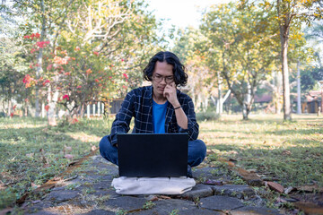 A male student wearing a plaid shirt and glasses sits in the park with his laptop. with a thoughtful expression.