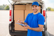 © Home-stock - Man courier in blue uniform holding clipboard and smiling at camera, checking list of clients, standing near van