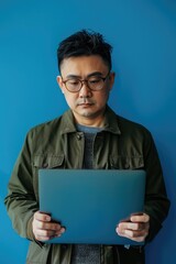 A smiling man in a green jacket and glasses, holding a light blue tablet computer.