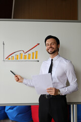 Wall Mural - A man is standing in front of a white board with a graph on it. He is holding a piece of paper and pointing to the graph. The man is smiling and he is giving a presentation