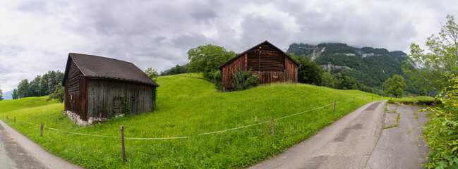 Panorama with old farmhouse on steep slope and rocky mountain in background. green pasture and forest with single tree in Rhein valley under Alpstein. peak with cloud hut, interesting cloudy day