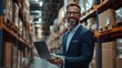 © liliyabatyrova - A cheerful businessman reviews inventory on his laptop amidst shelves of products in a bustling warehouse