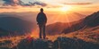 © StockUp - A person stands atop a mountain peak looking out towards the setting sun, surrounded by rolling hills and grassy plains.