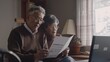 © VK Studio - An elderly couple, sitting together at home, carefully reviews a document near a laptop, bathed in gentle daylight through a window.