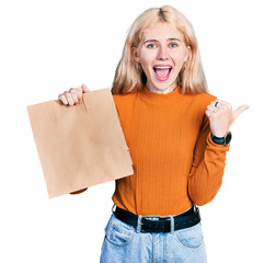 Canvas Print - Young caucasian woman holding take away paper bag pointing thumb up to the side smiling happy with open mouth