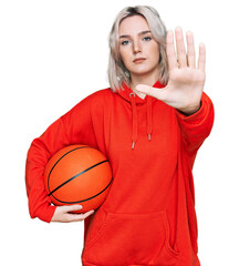 Poster - Young blonde girl holding basketball ball with open hand doing stop sign with serious and confident expression, defense gesture