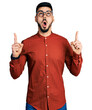 © Krakenimages.com - Young hispanic man with beard wearing business shirt and glasses amazed and surprised looking up and pointing with fingers and raised arms.