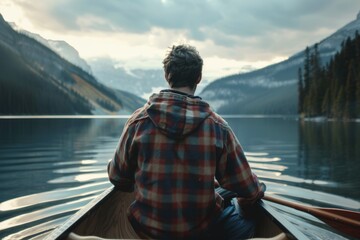 young man wearing a rugged flannel jacket and jeans sitting in a canoe on lake looking at distant mountains in background