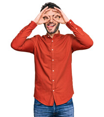 Poster - Young hispanic man wearing casual clothes doing ok gesture like binoculars sticking tongue out, eyes looking through fingers. crazy expression.