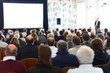 © 为轩 张 - Large audience in an auditorium listening to a speaker on stage with a white screen and light background, viewed from behind, ideal for business presentations or conferences.