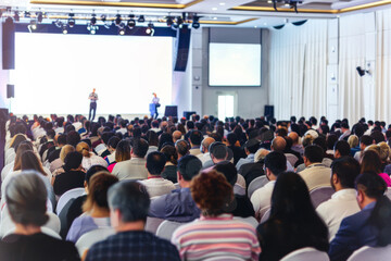 Large audience in an auditorium listening to a speaker on stage with a white screen and light background, viewed from behind, ideal for business presentations or conferences.