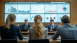 © Anastasia - A team of office workers in formal suits, sitting in a conference room with a large screen displaying their sales figures and growth statistics.