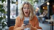 © Lens Legacy - A young blonde woman with freckles and an orange cardigan is holding a smartphone and showing a wide-eyed expression of surprise while seated in a trendy café.